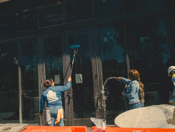 Three individuals engaged in cleaning large glass doors. One person is using a broom or mop to scrub the glass, another is spraying water, and the third person is wearing a hard hat and a safety vest. There are construction materials in the foreground.