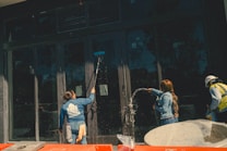 Three individuals engaged in cleaning large glass doors. One person is using a broom or mop to scrub the glass, another is spraying water, and the third person is wearing a hard hat and a safety vest. There are construction materials in the foreground.