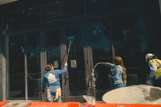 Three individuals engaged in cleaning large glass doors. One person is using a broom or mop to scrub the glass, another is spraying water, and the third person is wearing a hard hat and a safety vest. There are construction materials in the foreground.