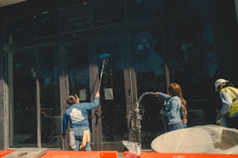Three individuals engaged in cleaning large glass doors. One person is using a broom or mop to scrub the glass, another is spraying water, and the third person is wearing a hard hat and a safety vest. There are construction materials in the foreground.