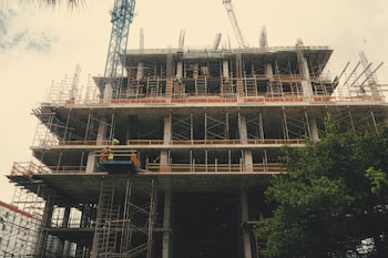 A construction site featuring a multi-story building in progress, surrounded by scaffolding and cranes. Workers in safety gear are visible on the site. The structure is partially completed with exposed concrete and metal frameworks.