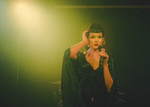 A person stands on stage holding a microphone with one hand while the other hand is raised to their head. The lighting is dramatic, casting a dark and moody ambiance. The person is wearing a dark, flowing outfit and has bold, striking makeup.
