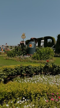 A beautifully landscaped garden with a variety of blooming flowers in the foreground. Central to the scene is an artistic display featuring a large stone structure with a trident on top, likely representing a Hindu deity. In the background, there are people, possibly statues, dressed in traditional clothing.
