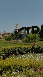 A beautifully landscaped garden with a variety of blooming flowers in the foreground. Central to the scene is an artistic display featuring a large stone structure with a trident on top, likely representing a Hindu deity. In the background, there are people, possibly statues, dressed in traditional clothing.