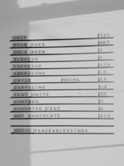 A monochrome menu board displays a list of various coffee and tea options, each paired with prices to the right. The items include drip, pour over, cold brew, Turkish, espresso, Americano, latte, mocha, cappuccino, flat white, cortado, assorted teas, and hot chocolate. A line at the bottom mentions the WiFi password 'peace&blessings'.