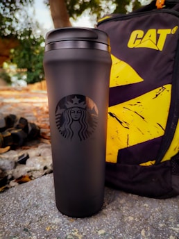 A sleek black travel mug with the Starbucks logo is placed on the ground next to a rugged backpack bearing the CAT logo. The background features a mix of concrete and fallen leaves, with hints of greenery.