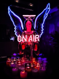 A neon-lit angel figure with glowing blue wings and a pink halo holds a sign that reads 'ON AIR.' Below, a collection of lit candles in various colors adds to the vibrant ambiance against a dark background.