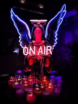 A neon-lit angel figure with glowing blue wings and a pink halo holds a sign that reads 'ON AIR.' Below, a collection of lit candles in various colors adds to the vibrant ambiance against a dark background.