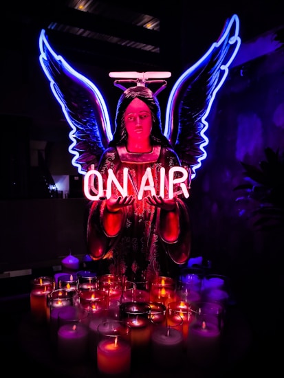 A neon-lit angel figure with glowing blue wings and a pink halo holds a sign that reads 'ON AIR.' Below, a collection of lit candles in various colors adds to the vibrant ambiance against a dark background.