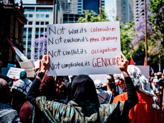 A crowd gathers in a city street holding signs with bold messages criticizing war, eviction, conflict, and complicated policies, framing them as colonialism, ethnic cleansing, occupation, and genocide. The background features urban buildings and trees in bloom.