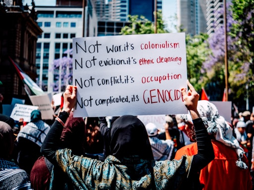 A crowd gathers in a city street holding signs with bold messages criticizing war, eviction, conflict, and complicated policies, framing them as colonialism, ethnic cleansing, occupation, and genocide. The background features urban buildings and trees in bloom.