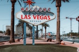 A famous 'Welcome to Fabulous Las Vegas, Nevada' sign stands prominently in front of a backdrop of palm trees and high-rise buildings. The sign is adorned with retro-style, vibrant red and blue lettering, and is situated on a neatly manicured island surrounded by a road with passing cars. Palm trees add a tropical feel to the scene, complemented by clear blue skies.