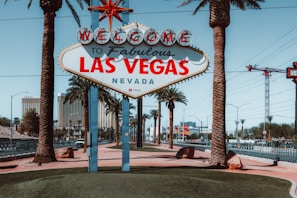 A famous 'Welcome to Fabulous Las Vegas, Nevada' sign stands prominently in front of a backdrop of palm trees and high-rise buildings. The sign is adorned with retro-style, vibrant red and blue lettering, and is situated on a neatly manicured island surrounded by a road with passing cars. Palm trees add a tropical feel to the scene, complemented by clear blue skies.