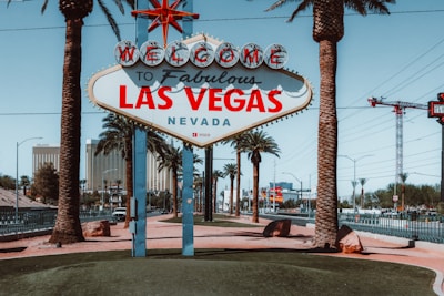 A famous 'Welcome to Fabulous Las Vegas, Nevada' sign stands prominently in front of a backdrop of palm trees and high-rise buildings. The sign is adorned with retro-style, vibrant red and blue lettering, and is situated on a neatly manicured island surrounded by a road with passing cars. Palm trees add a tropical feel to the scene, complemented by clear blue skies.