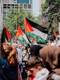 A large group of people is gathered, waving Palestinian flags. They are outdoors in an urban area with modern buildings and leafy trees in the background. The crowd comprises of individuals from various age groups and genders, some wearing headscarves and traditional attire.