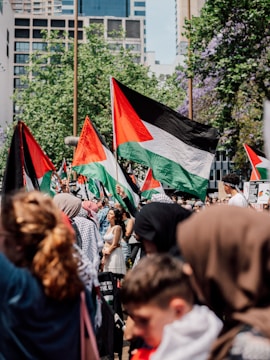 A large group of people is gathered, waving Palestinian flags. They are outdoors in an urban area with modern buildings and leafy trees in the background. The crowd comprises of individuals from various age groups and genders, some wearing headscarves and traditional attire.