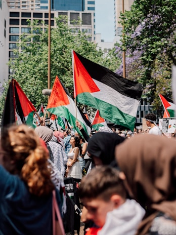 A large group of people is gathered, waving Palestinian flags. They are outdoors in an urban area with modern buildings and leafy trees in the background. The crowd comprises of individuals from various age groups and genders, some wearing headscarves and traditional attire.