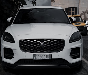 A sleek white SUV is parked with a prominent grille and modern headlights. The car is in a parking area with a couple of other vehicles visible in the background, including a yellow and black van. The setting appears to be an outdoor urban environment, with part of a building visible in the background.