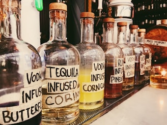 Several glass bottles with cork stoppers are lined up on a counter. Each bottle has a handwritten label indicating the type of alcohol and the infusion flavor such as 'Vodka Infused Butterfly' and 'Tequila Infused Corn.' The setting appears to be in a bar or restaurant with other bottles and bar tools in the background.