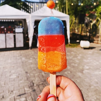 A person is holding a colorful popsicle with distinct blue, red, and orange layers. In the background, there is an outdoor setting with two white tents and some blurred greenery and lights.