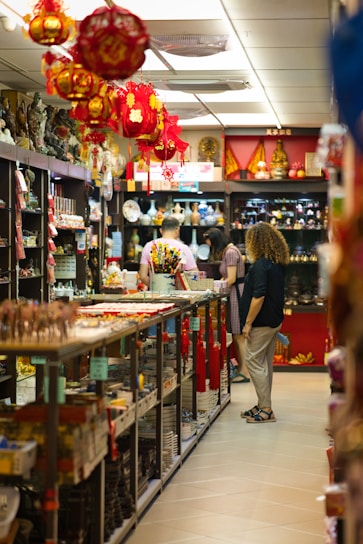 A store aisle filled with various items, including red hanging lanterns, shelves stocked with ceramics, souvenirs, and decorative objects. Several people are browsing the merchandise. The store is brightly lit with a warm ambiance.