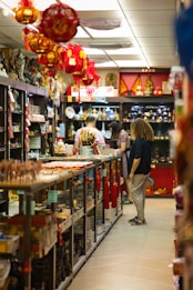 A store aisle filled with various items, including red hanging lanterns, shelves stocked with ceramics, souvenirs, and decorative objects. Several people are browsing the merchandise. The store is brightly lit with a warm ambiance.