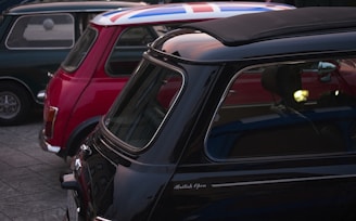 A group of classic Mini Cooper cars parked closely together. The car in the foreground is black with a partial soft top open and has the words 'British Open' inscribed on it. The car behind it is red with a roof featuring the design of the Union Jack. A green Mini Cooper is also visible, parked alongside.