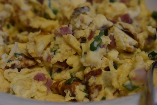 A close-up view of scrambled eggs mixed with pieces of bacon and green herbs. The eggs are soft and fluffy, with a slightly golden texture and visible browned edges. The bacon is finely chopped, adding a pinkish hue and savory appearance. Green herbs are scattered throughout, adding a fresh touch.