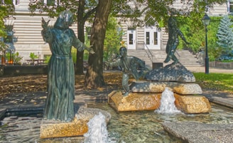An outdoor statue installation featuring three bronze figures in dynamic poses situated in a small water fountain structure. The scene is surrounded by lush greenery, and a classic building facade is visible in the background. The statues depict a woman in a long robe and two children in playful postures, all positioned on stone slabs above the bubbling water.