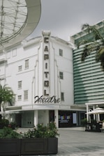 A white building labeled with 'Capitol Theatre' in vintage-style lettering. The facade includes multiple windows and decorative elements. Nearby, there are plants, a palm tree, and modern structures with horizontal lines. A broad glass canopy is partially visible near the top left corner. Below, there are signs for 'Wonderland' exhibit and an outdoor seating area with umbrellas.