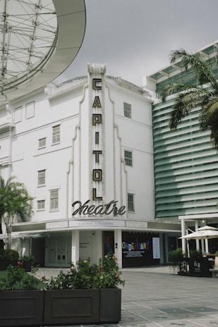 A white building labeled with 'Capitol Theatre' in vintage-style lettering. The facade includes multiple windows and decorative elements. Nearby, there are plants, a palm tree, and modern structures with horizontal lines. A broad glass canopy is partially visible near the top left corner. Below, there are signs for 'Wonderland' exhibit and an outdoor seating area with umbrellas.