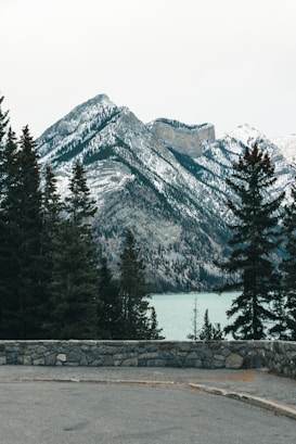 A serene landscape featuring snow-capped mountains in the background, with a calm, turquoise lake nestled at their base. Tall evergreen trees frame the scene, and a stone wall borders a paved area in the foreground. The overall atmosphere is peaceful and natural.