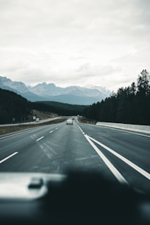 An open highway stretches into the distance, flanked by tall pine trees and leading towards majestic mountains under a cloudy sky. A few cars are visible on the road, adding to the sense of travel and journey.