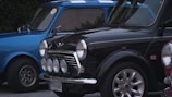 Two classic Mini Cooper cars are parked side by side, featuring a blue and black one. Both cars have iconic front grilles and circular headlights, with the black car displaying additional fog lights. The Union Jack emblem is present on the black car, and vintage design elements are evident.
