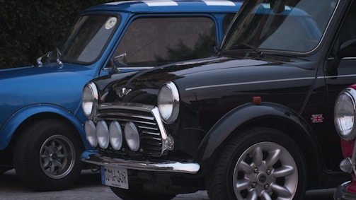 Two classic Mini Cooper cars are parked side by side, featuring a blue and black one. Both cars have iconic front grilles and circular headlights, with the black car displaying additional fog lights. The Union Jack emblem is present on the black car, and vintage design elements are evident.