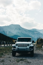 A white SUV is parked on a gravel path near a road with a scenic backdrop of towering mountains and a partly cloudy sky. The landscape features lush green trees and vegetation, creating a serene and adventurous atmosphere.