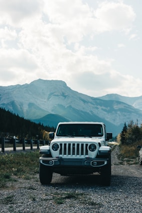 A white SUV is parked on a gravel path near a road with a scenic backdrop of towering mountains and a partly cloudy sky. The landscape features lush green trees and vegetation, creating a serene and adventurous atmosphere.