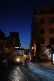 A classic yellow tram travels along cobbled streets under a deep blue evening sky. Shadows from buildings and soft street lighting create a warm ambiance. The tram, labeled 'M. Moniz', highlights historic transportation amidst an urban setting. A few people can be seen through the tram windows, and cars are visible further down the street.