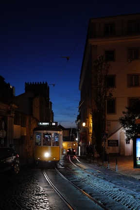 A classic yellow tram travels along cobbled streets under a deep blue evening sky. Shadows from buildings and soft street lighting create a warm ambiance. The tram, labeled 'M. Moniz', highlights historic transportation amidst an urban setting. A few people can be seen through the tram windows, and cars are visible further down the street.