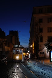 A classic yellow tram travels along cobbled streets under a deep blue evening sky. Shadows from buildings and soft street lighting create a warm ambiance. The tram, labeled 'M. Moniz', highlights historic transportation amidst an urban setting. A few people can be seen through the tram windows, and cars are visible further down the street.