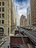 A cityscape featuring a modern urban environment with a mix of historic and contemporary architecture. The central focus is a tall skyscraper in the background, surrounded by other mid-rise buildings. There is an elevated train track in the foreground with a train traveling along it. Pedestrians and vehicles are present on the street below.