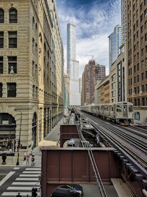 A cityscape featuring a modern urban environment with a mix of historic and contemporary architecture. The central focus is a tall skyscraper in the background, surrounded by other mid-rise buildings. There is an elevated train track in the foreground with a train traveling along it. Pedestrians and vehicles are present on the street below.