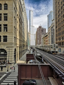 A cityscape featuring a modern urban environment with a mix of historic and contemporary architecture. The central focus is a tall skyscraper in the background, surrounded by other mid-rise buildings. There is an elevated train track in the foreground with a train traveling along it. Pedestrians and vehicles are present on the street below.