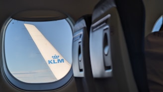 An airplane wing with the KLM logo is visible through a passenger window, alongside an empty row of seats inside the aircraft. The sky appears clear with a few clouds.