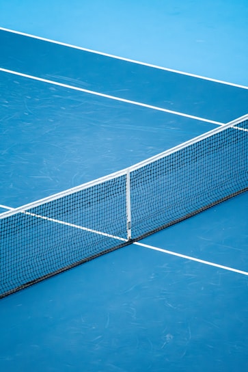 A blue tennis court featuring a central black net with white lines marking the playing areas.