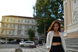 A woman is standing confidently in an urban setting. She is wearing a white blazer over a black top and pants, accessorized with gold jewelry. In the background, there is an elegant old building with multiple windows, and parked cars can be seen on the street.