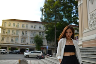 A woman is standing confidently in an urban setting. She is wearing a white blazer over a black top and pants, accessorized with gold jewelry. In the background, there is an elegant old building with multiple windows, and parked cars can be seen on the street.