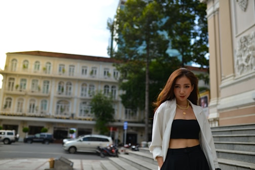 A woman is standing confidently in an urban setting. She is wearing a white blazer over a black top and pants, accessorized with gold jewelry. In the background, there is an elegant old building with multiple windows, and parked cars can be seen on the street.