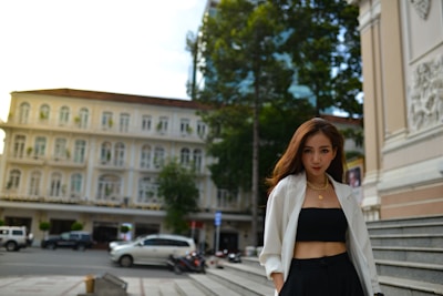 A woman is standing confidently in an urban setting. She is wearing a white blazer over a black top and pants, accessorized with gold jewelry. In the background, there is an elegant old building with multiple windows, and parked cars can be seen on the street.