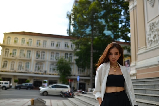 A woman is standing confidently in an urban setting. She is wearing a white blazer over a black top and pants, accessorized with gold jewelry. In the background, there is an elegant old building with multiple windows, and parked cars can be seen on the street.